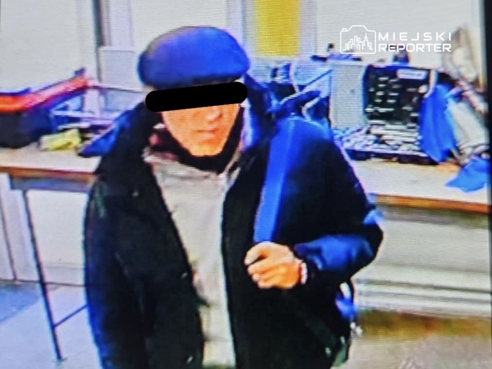 Mężczyzna w czapce i ciemnej kurtce trzyma torbę, stojąc w pomieszczeniu z komputerami i sprzętem elektronicznym.