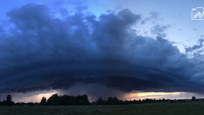 Ciemne chmury burzowe gromadzą się na horyzoncie, nad zieloną łąką i drzewami, w tle widać zachodzące słońce.