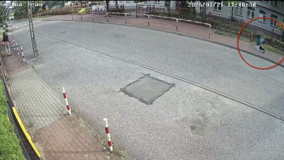 Mężczyzna w ciemnej odzieży spacerujący po chodniku przy ulicy, obok znaku drogowym i ogrodzeniu.