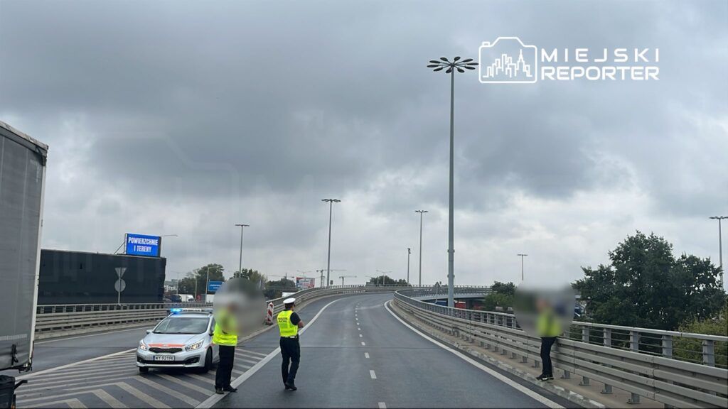 Policjanci w kamizelkach odblaskowych kierują ruchem na drodze, obok zaparkowany radiowóz z włączonymi światłami.