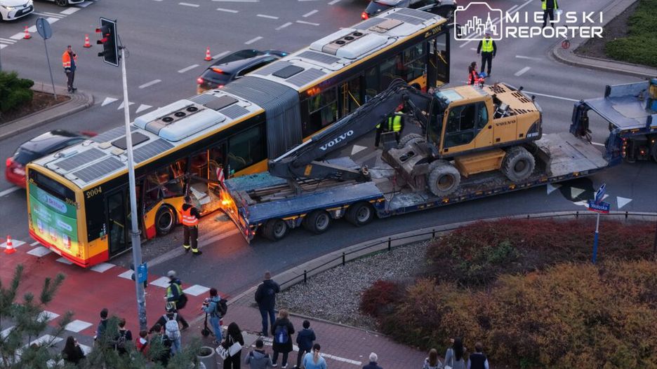 Autobus z uszkodzoną tylną częścią jest holowany przez ciężki sprzęt, a obok gromadzi się grupa gapiów.