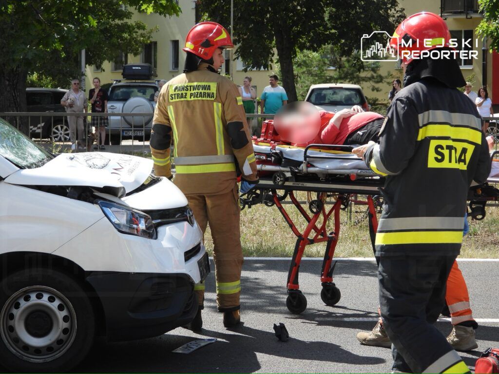 Strażacy transportują poszkodowanego na noszach obok uszkodzonego białego samochodu na drodze. W tle obserwatorzy.