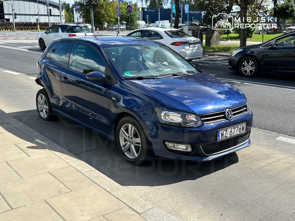 Niebieski samochód osobowy marki Volkswagen z uszkodzeniem bocznym stoi na chodniku obok ruchliwej ulicy.