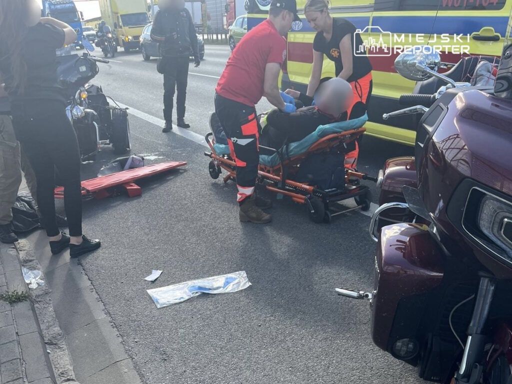 Ratownicy medyczni udzielają pomocy poszkodowanemu na jezdni, obok przewróconego motocykla. W tle przejeżdżające pojazdy.