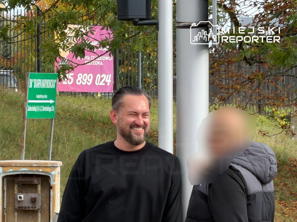 Mężczyzna w czarnej bluzie uśmiecha się, stojąc obok drugiej osoby w kurtce, w otoczeniu znaków drogowych.