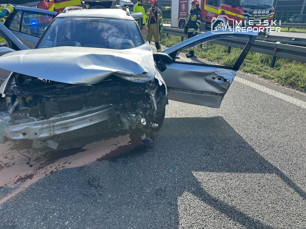 W wyniku wypadku drogowego uszkodzone zostało auto osobowe, które zatrzymało się na poboczu. Widoczne są zniszczenia karoserii oraz otwarte drzwi. Na miejscu zdarzenia pracują służby ratunkowe, w tym straż pożarna, zajmujące się zabezpieczeniem terenu.