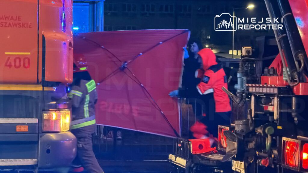 Ratownicy w odzieży ochronnej ustawiają czerwoną osłonę obok wozu strażackiego w nocy na ulicy.