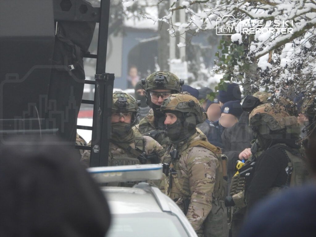 Grupa uzbrojonych funkcjonariuszy w kamizelkach taktycznych rozmawia obok pojazdu policyjnego w zaśnieżonym otoczeniu.