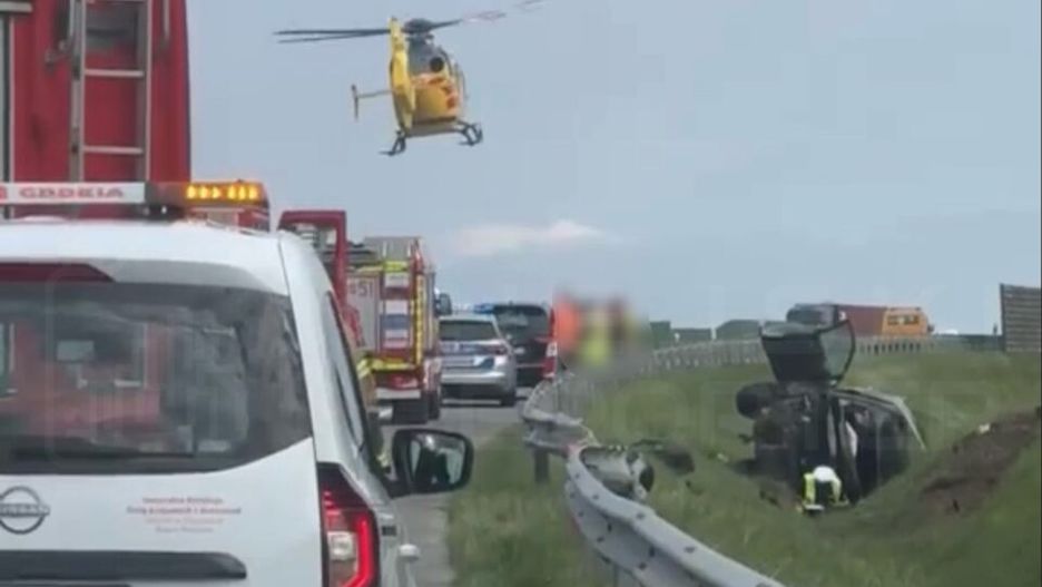 Żółty helikopter lądował obok drogi, gdzie przewrócone auto znajduje się w rowie, a pojazdy służb ratunkowych blokują pas.