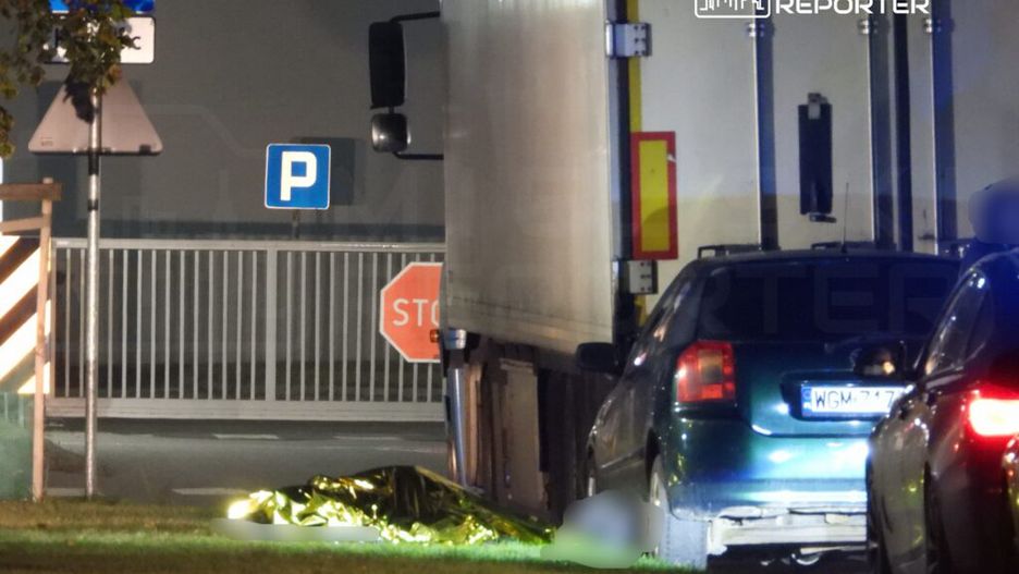 Złoty koc leży na trawie obok zaparkowanego samochodu, w tle widoczne znaki drogowe i ciężarówka.