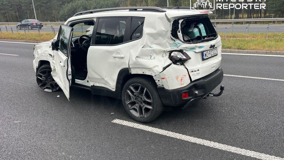 Uszkodzony biały SUV stoi na poboczu drogi, z otwartymi drzwiami i zniszczonym tyłem. W tle widać inne pojazdy.