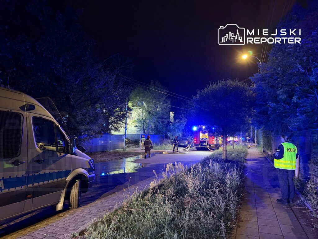 Na drodze w nocy stoją pojazdy służb ratunkowych, a strażacy rozkładają węże gaśnicze w otoczeniu oświetlonym niebieskimi światłami.