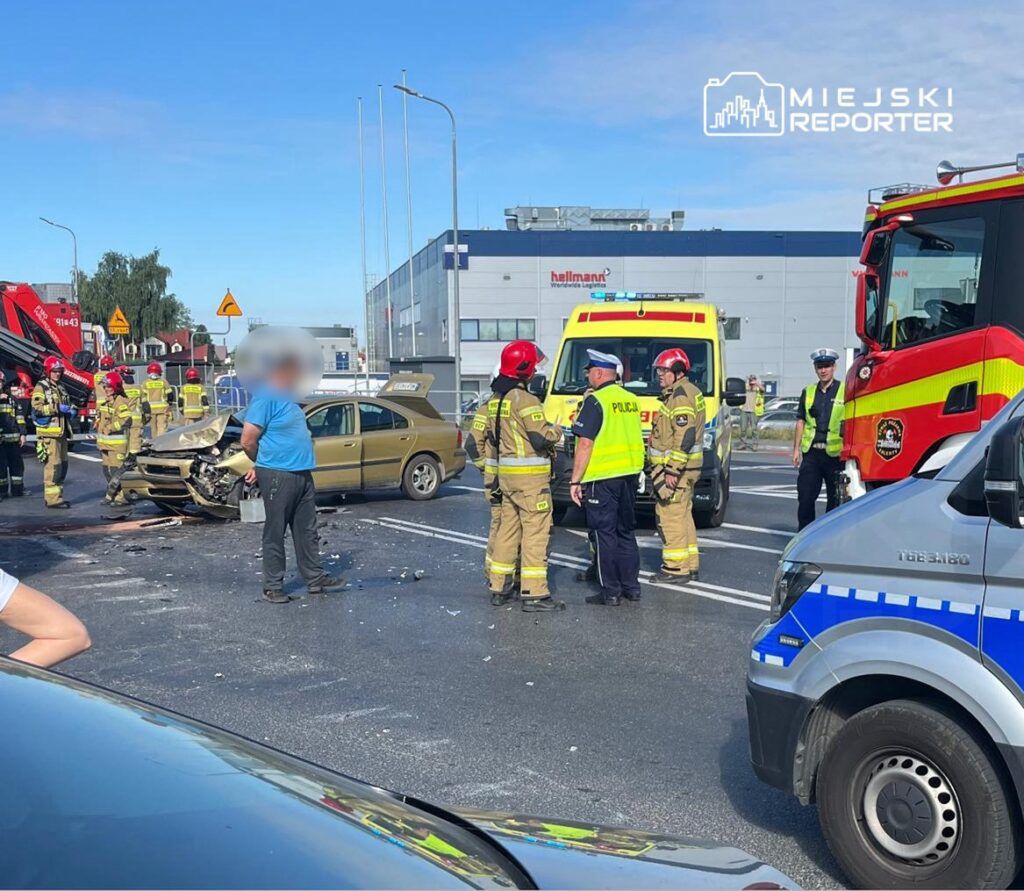 Grupa strażaków i policjantów prowadzi działania na miejscu wypadku drogowego, obok uszkodzonego samochodu.