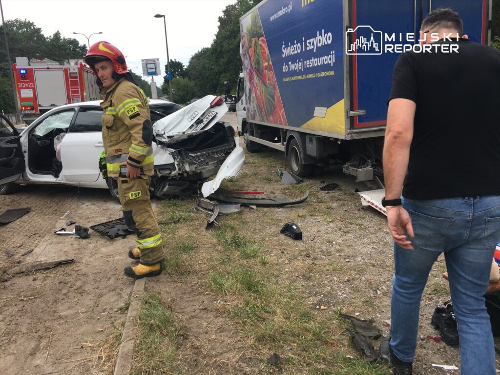 Strażak w mundurze stoi obok rozbitego samochodu osobowego, który zderzył się z ciężarówką na poboczu drogi.
