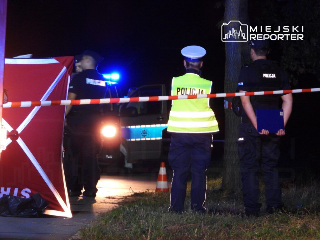 Policjanci zabezpieczają miejsce zdarzenia w nocy. Na pierwszym planie widoczny jest funkcjonariusz w kamizelce odblaskowej oraz dwóch policjantów, którzy rozmawiają. W tle radiowóz policyjny z włączonymi sygnałami świetlnymi. Obszar otoczony taśmą zabezpieczającą.