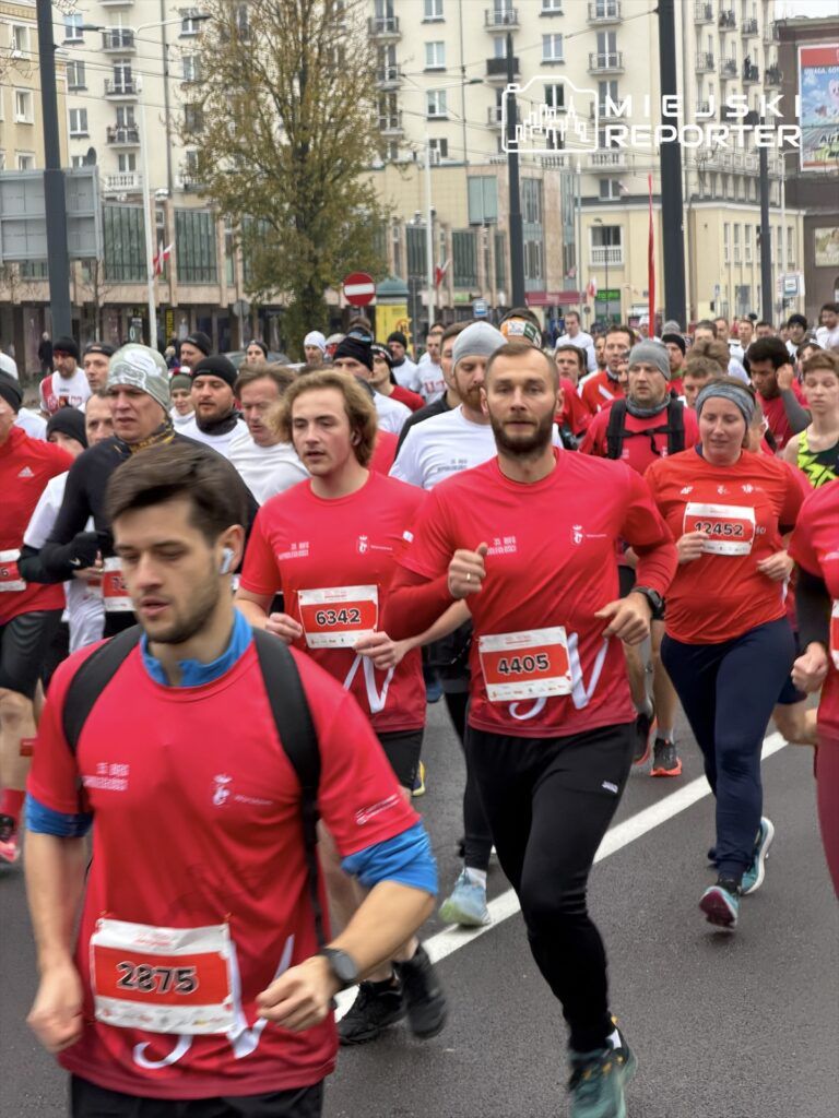 Grupa biegaczy w czerwonych koszulkach uczestniczy w wydarzeniu biegowym na miejskiej ulicy.