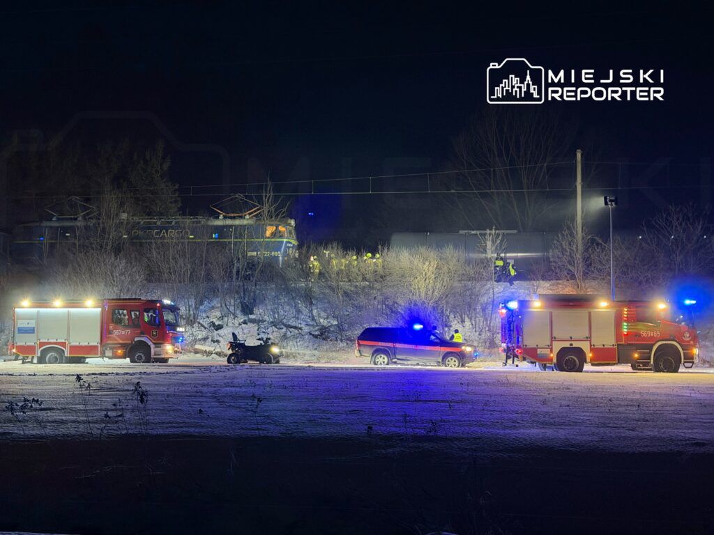 W nocy strażacy pracują nad usunięciem pojazdu z torów kolejowych, obok stoi wóz strażacki.