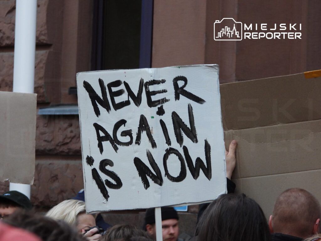 Osoba trzymająca transparent z napisem "NEVER AGAIN IS NOW" na tle tłumu podczas demonstracji.