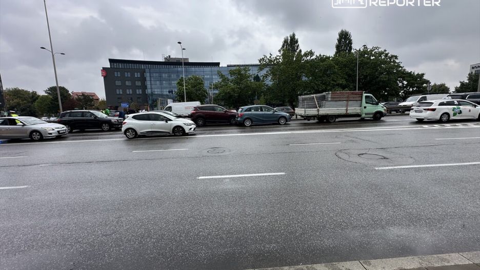 Ruch samochodowy na ulicy w deszczowy dzień, z różnymi pojazdami oraz nowoczesnym biurowcem w tle.