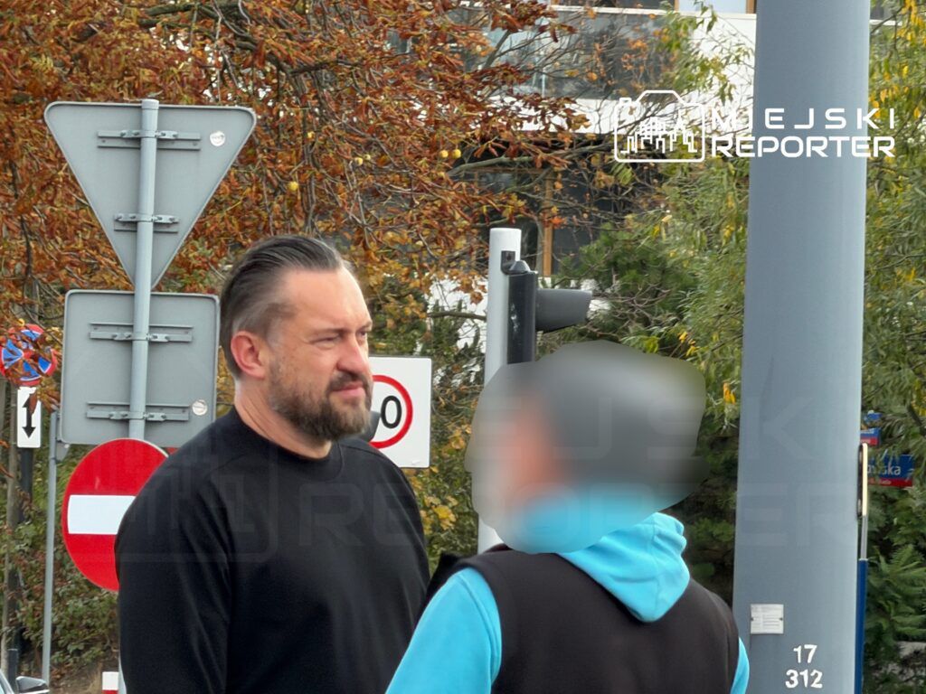 Mężczyzna z brodą w czarnej bluzie rozmawia z osobą w niebieskiej kurtce na ulicy, w tle znaki drogowe.
