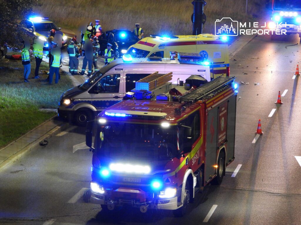 Grupa ratowników i policjantów pracuje przy wypadku drogowym, obok wozów strażackich i ambulansów.