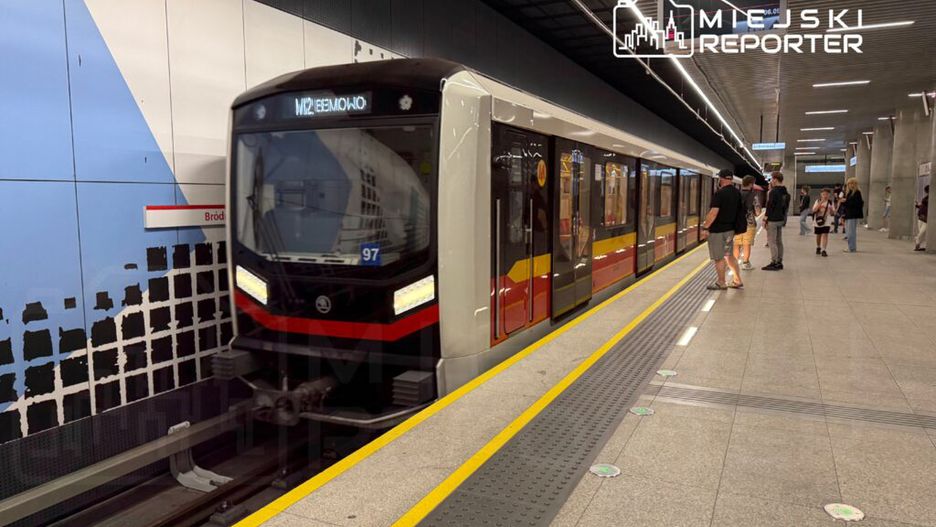 Pociąg metra wjeżdża na stację, a pasażerowie czekają na peronie w nowoczesnym otoczeniu.