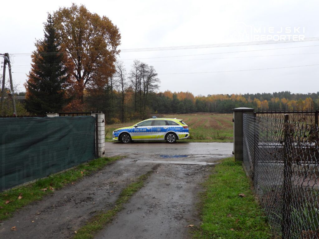 Policyjna radiowóz zaparkowany przy drodze w otoczeniu drzew oraz pól, w deszczowy dzień.
