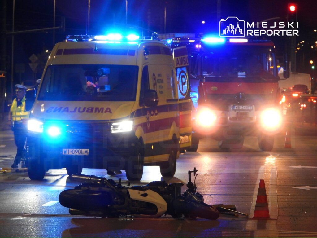Karetka pogotowia z włączonymi sygnałami świetlnymi na drodze obok przewróconego motocykla, w tle woz strażacki.