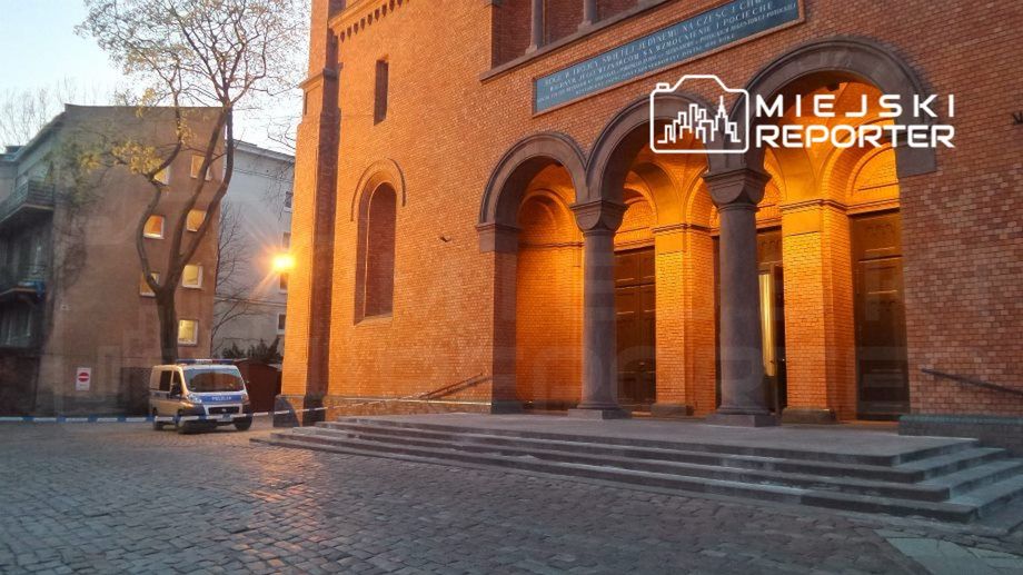 Mężczyzna w niebieskim vanie parkuje przed ceglaną budowlą z łukowymi wejściami oświetlonymi ciepłym światłem.