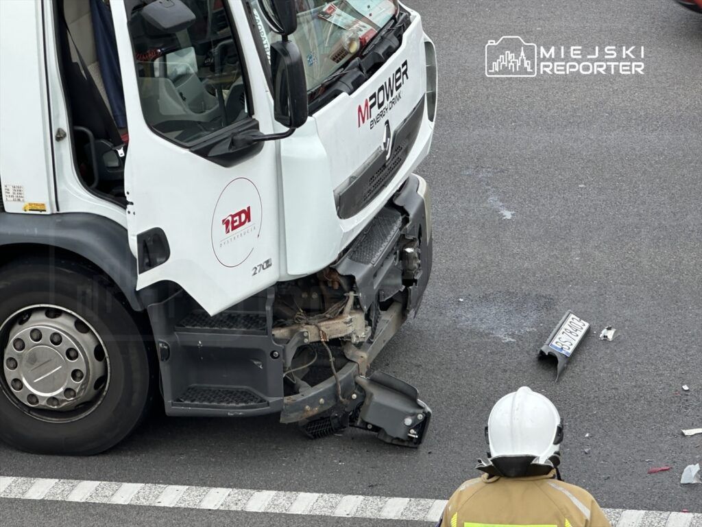 Wielka ciężarówka z uszkodzoną przednią częścią stoi na drodze, obok niej strażak w hełmie. Na jezdni leży fragment tablicy rejestracyjnej.