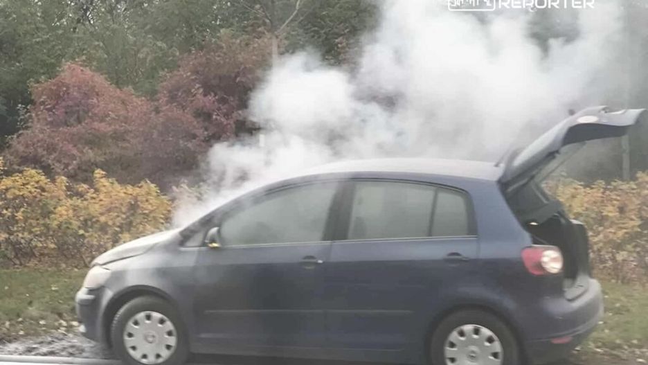 Niebieski samochód osobowy wydobywający dym, zaparkowany na poboczu drogi w otoczeniu drzew i krzewów.