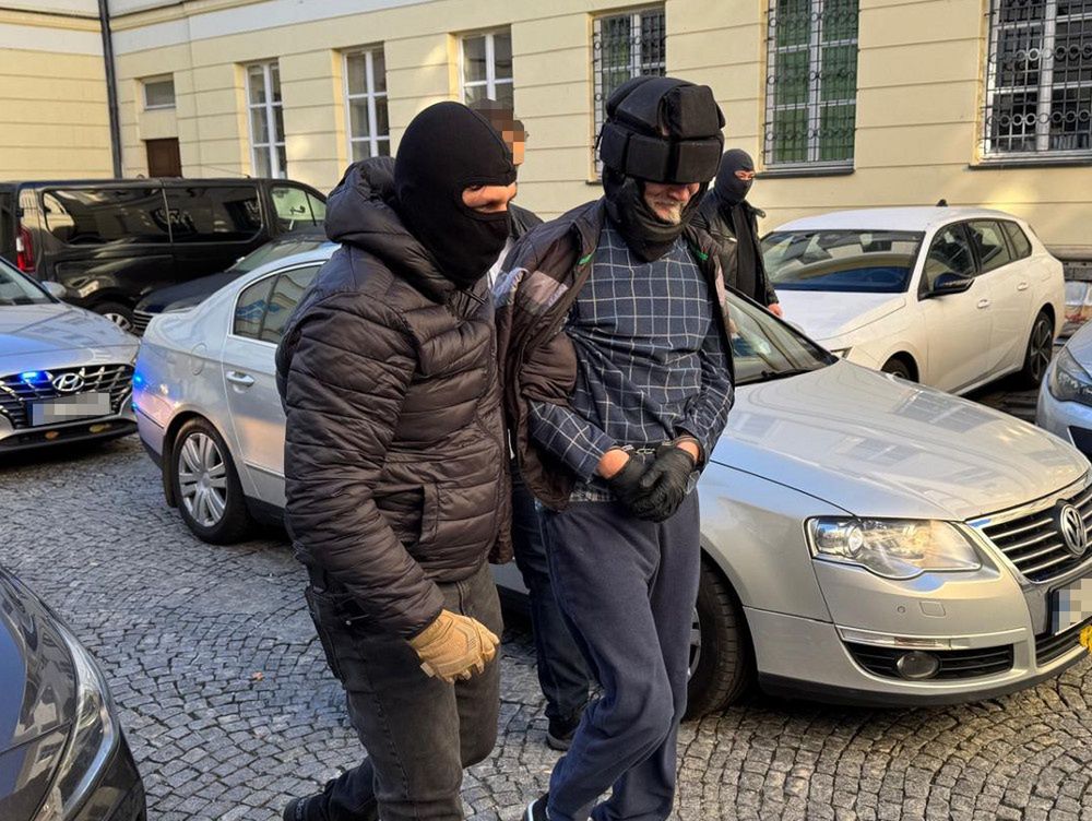 Mężczyzna w dresie prowadzony przez zamaskowanego funkcjonariusza wśród zaparkowanych samochodów.