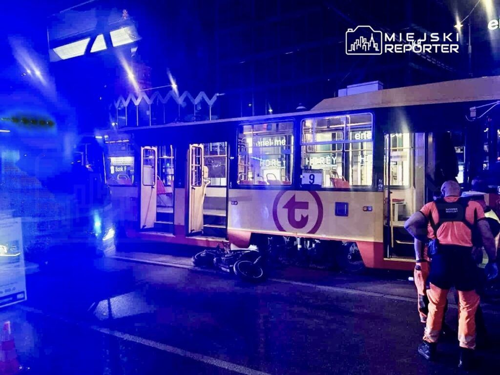 Na ulicy stoi tramwaj z otwartymi drzwiami, obok pracownicy w pomarańczowych kamizelkach zabezpieczają miejsce zdarzenia.