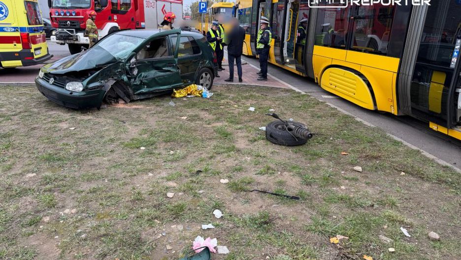 Zniszczony samochód osobowy stoi na trawie obok żółtego tramwaju, a strażacy i policjanci pomagają poszkodowanym.