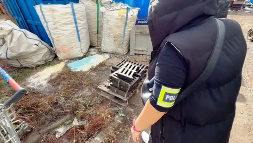 Funkcjonariusz policji bada teren w pobliżu worków z odpadami i paletą na zarośniętej trawie.