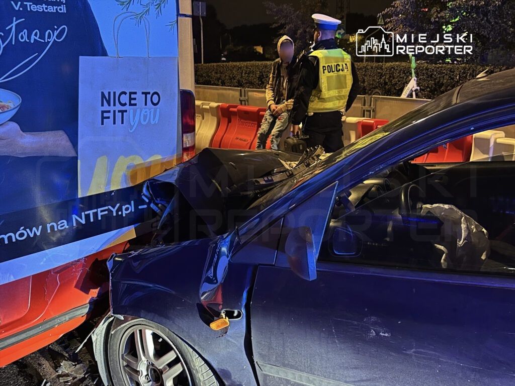 Zderzenie dwóch pojazdów w nocy, z policjantem rozmawiającym z osobą siedzącą na krawężniku obok uszkodzonego samochodu.