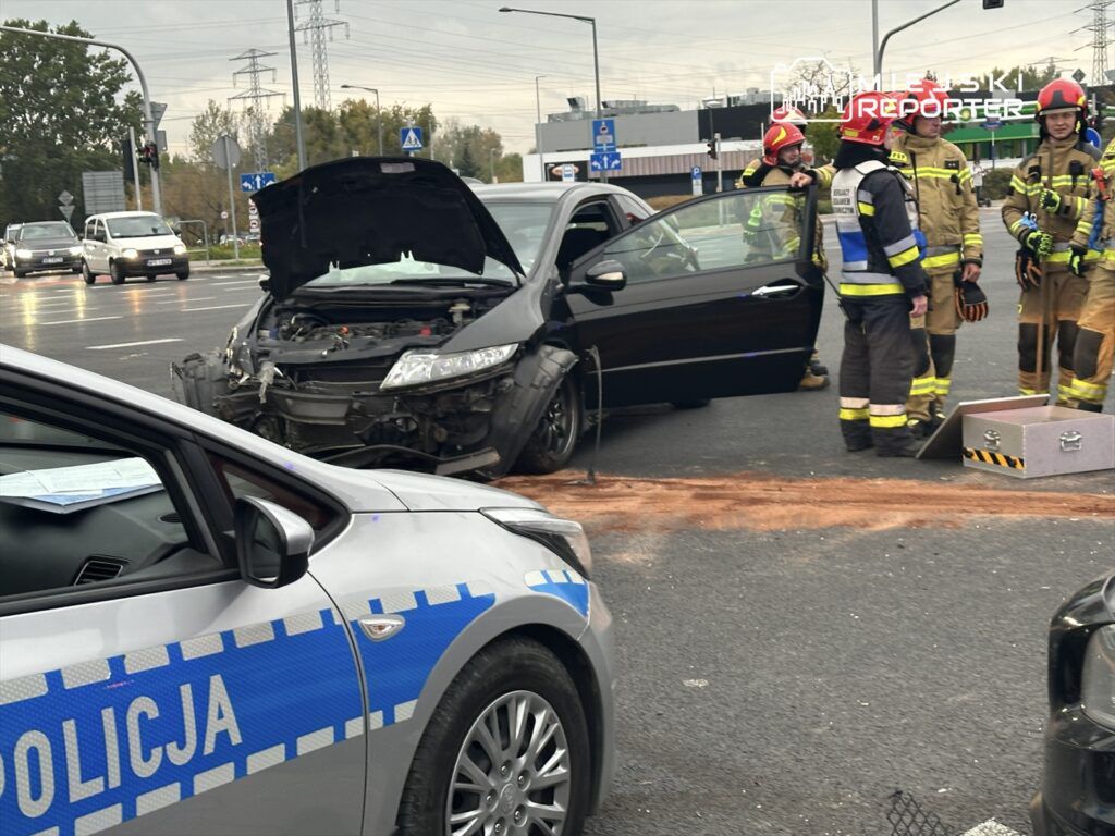 Czterech strażaków w mundurach rozmawia z policjantem przy uszkodzonym samochodzie na skrzyżowaniu.