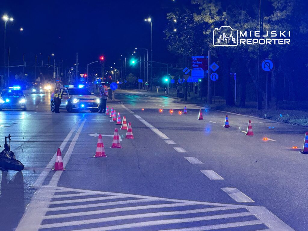 Policjanci z niebieskimi sygnałami kontrolują ruch na zamkniętej drodze, otoczonej pachołkami.