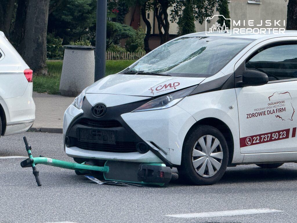 Zniszczony skuter elektryczny leży pod przednim zderzakiem białego samochodu osobowego na miejskiej ulicy.