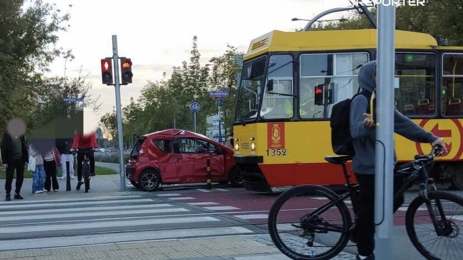 Czerwony samochód osobowy uszkodzony po zderzeniu z tramwajem na skrzyżowaniu, obok przechodnie i rowerzysta.
