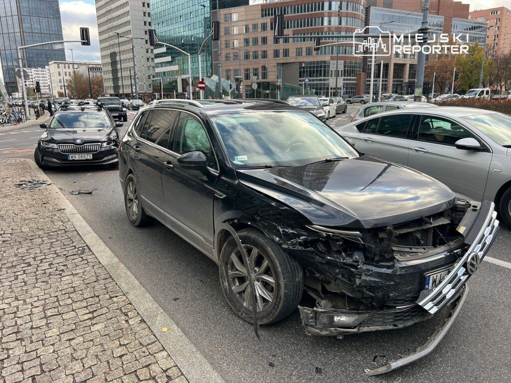 Czarny samochód osobowy z uszkodzonym przodem zaparkowany na ruchliwej ulicy w otoczeniu nowoczesnych budynków.