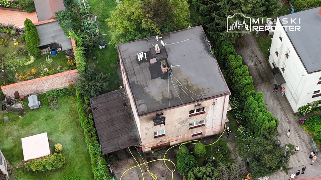 Dwupiętrowy budynek z uszkodzoną elewacją, otoczony zielenią, z wężami strażackimi na ziemi i ludźmi w pobliżu.