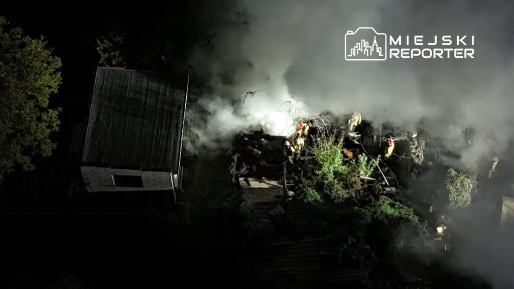 Strażacy gaszą pożar w zaroślach obok drewnianego budynku, otoczeni gęstym dymem w nocy.