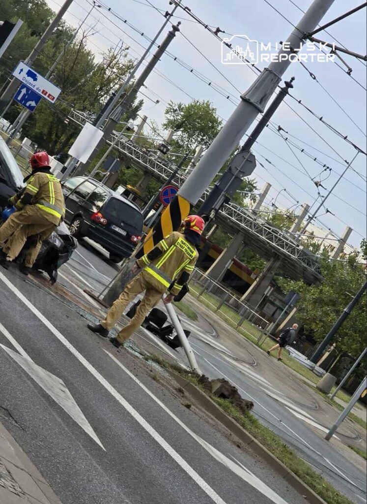 Dwóch strażaków w mundurach zajmuje się usuwaniem skutków wypadku drogowego przy zniszczonym słupie.