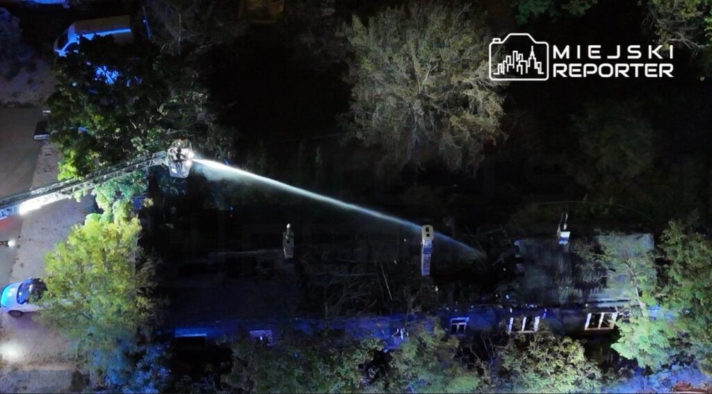 Strażacy używają węża gaśniczego z podnośnika, aby gasić pożar w zniszczonym budynku otoczonym drzewami.