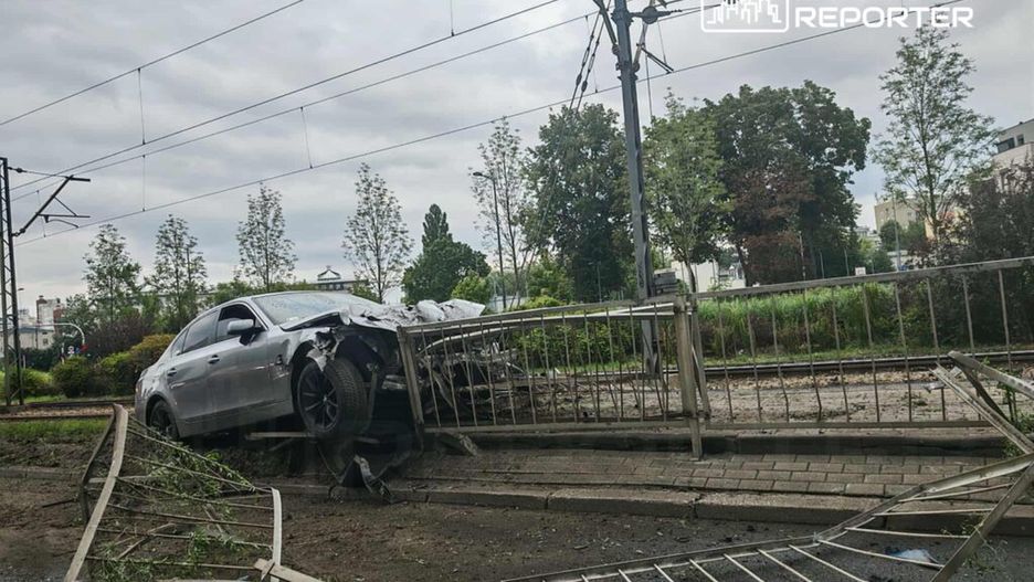 Uszkodzony samochód osobowy unieruchomiony na ogrodzeniu w pobliżu torów kolejowych, z widocznymi uszkodzeniami.