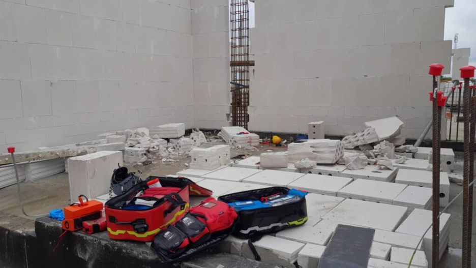 Na placu budowy leżą fragmenty białych bloczków, a obok znajdują się torby z narzędziami i sprzętem budowlanym.