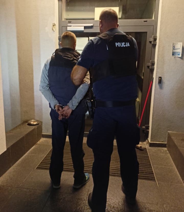 Mężczyzna z rękami skuci za plecami stoi przed wejściem do budynku, obok niego policjant w mundurze.