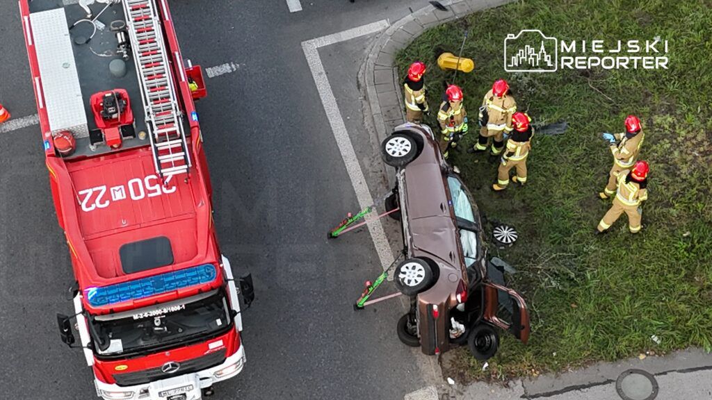 Ratownicy w mundurach pracują przy przewróconym samochodzie osobowym na skrzyżowaniu, obok wozu strażackiego.
