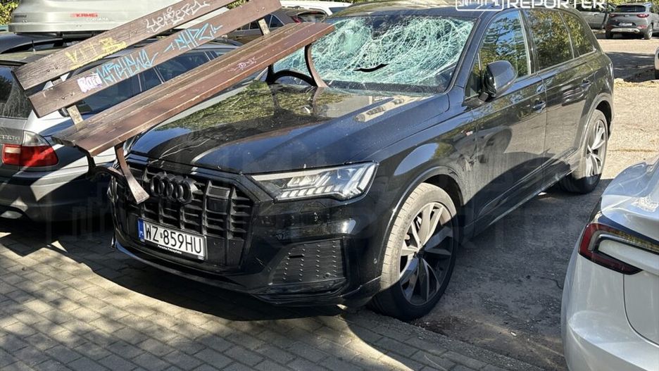 Czarny SUV z uszkodzoną szybą i drewnianą ławką unieruchomioną na dachu, zaparkowany między innymi pojazdami.